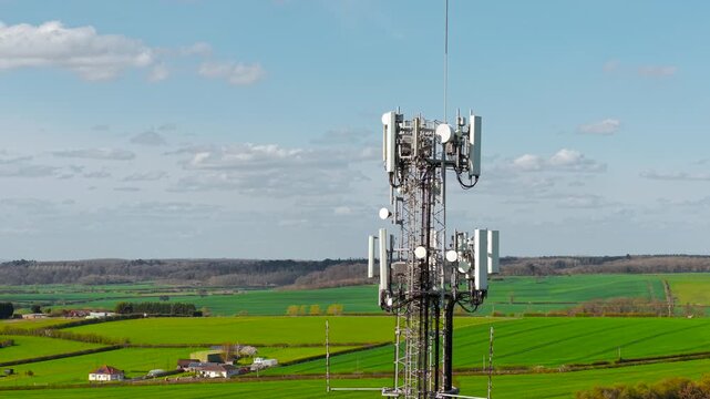 High cellular tower with 4G 5G antennas standing over agricultural fields under blue sky. Communication infrastructure with rural terrain and broadband network access. Telecom network coverage and