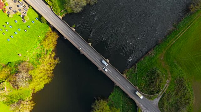 Diagonal aerial view of vehicles including compact car and small van crossing bridge over dark river with ripples and grassy fields. Rural road network with bridge connection and natural landscape
