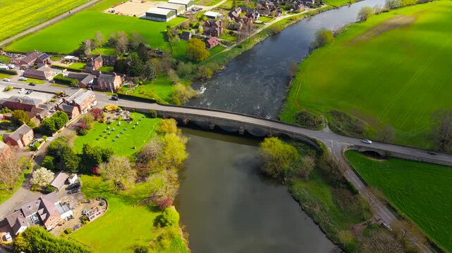 High altitude aerial of village bridge with vehicles including van and compact car moving between rural houses and open fields along river. Regional road passing through countryside with river