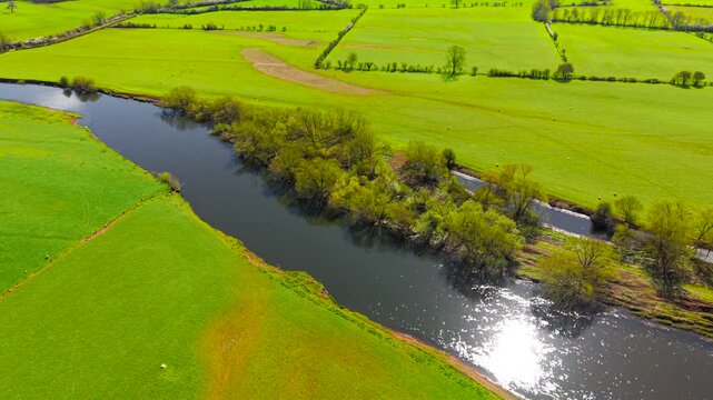 Close aerial view river channel reflecting sunlight surrounded by grassy fields and dense riverside trees. Detailed top view textures water surface vegetation and farmland borders. Natural habitat