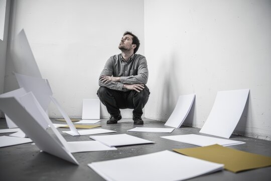 Man squatting among scattered papers and boards in a minimal studio space