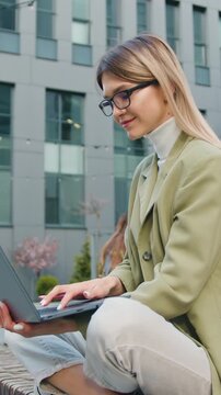 Attractive young caucasian female working or studying using laptop while sitting on bench of campus lawn. Pretty girl student preparing for classes in outdoor. Slow motion
