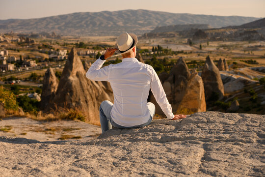 Young man in straw hat and white shirt sitting on a rock overlooking Cappadocia valley in Turkey. Inspiring travel experience, important life memories, freedom of travel, and wellness lifestyle