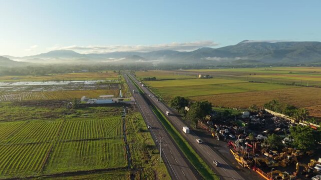 High-angle drone shot of Duarte Highway with vehicle traffic through green rice plantations. Scenic rural landscape with misty mountains and morning light in Dominican Republic