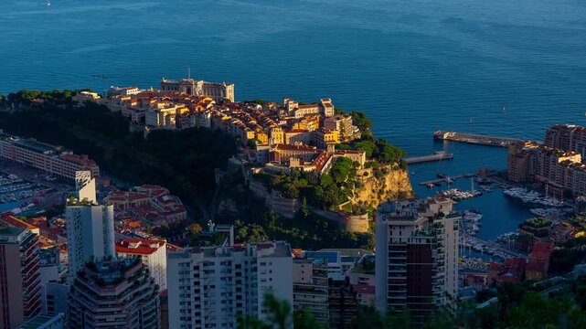 High-angle sunset timelapse of Monaco-Ville on the rock, flanked by Port Hercule and Fontvieille. Features sunlight fading into deep dusk over the Mediterranean.