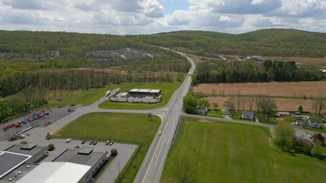 A high-angle drone shot overlooks a paved road junction and a modern commercial building, with expansive green fields and rural scenery in the background.