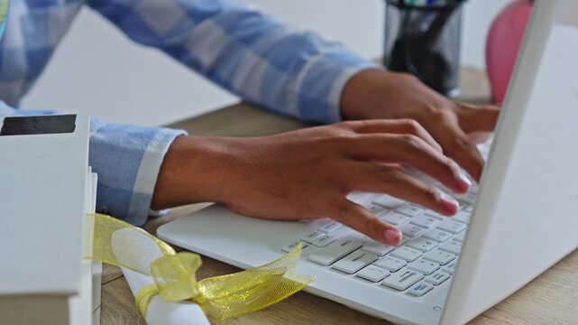 Close-up of student typing on laptop with diploma and books on desk