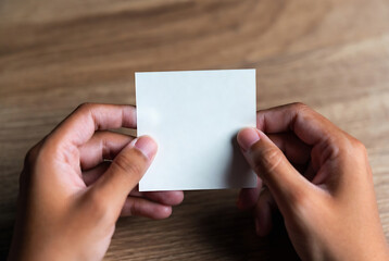 Obraz premium Hands are holding a blank white square of paper. It’s on a wooden table.
