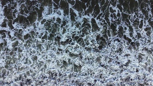 The ocean waves roll and crash against the sand, creating frothy white foam at the shore. The sun shines bright, making the water appear clear and blue, showing movement and energy.