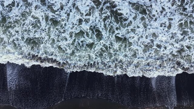 Waves roll onto the sandy shore, mixing foam with water. The day shows clear signs of movement as the ocean meets the land, creating patterns in the foam.
