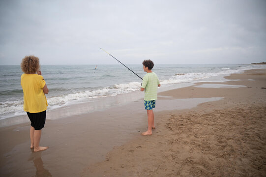 Middle aged woman watching her teenage son surf fishing on a quiet beach during summer vacation, standing barefoot by the shoreline under an overcast coastal sky