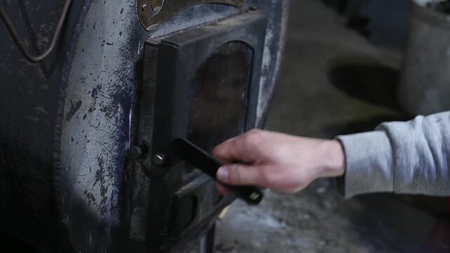 A man throws wood into a solid fuel boiler, close-up of his hands. He opens the stove door and loads the wood into the firebox.