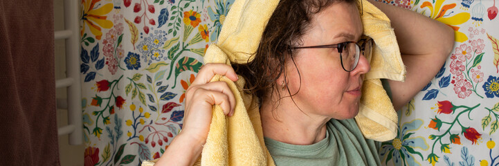 A woman performs her morning bathroom routine. She stands with a towel on her head, looking in the mirror. The colorful floral wall adds brightness to the space, banner © pundapanda