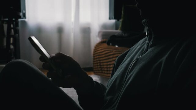 Person sits in silhouette while looking at a glowing mobile phone screen. Bright light from the device illuminates the hand in a dark indoor room. Daylight filters through curtains.