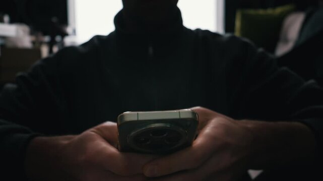 Man hands hold a smartphone in a dimly lit room. The screen light illuminates his fingers as he interacts with the device. Dark shadows create a moody and private atmosphere.
