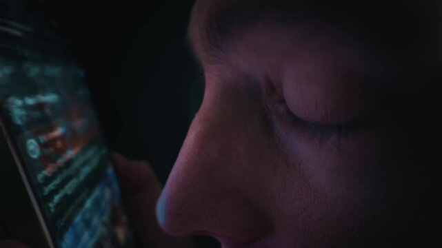 A close-up of a man staring at a smartphone screen in a completely dark room. The bright device light illuminates his face and reflects in his eye. He scrolls through digital media.