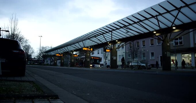 Ground level view of a bus station terminal with digital signs and glass awnings at dusk.