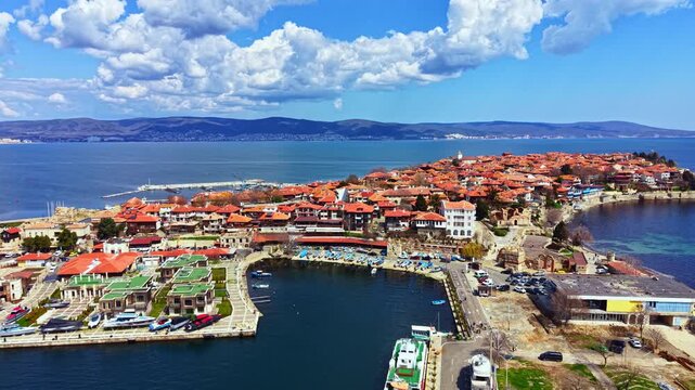 Nesebar Old Town shows a view of the coast with buildings and boats. The scene includes the harbor alongside the seaside and rolling hills in the background under a cloudy sky.