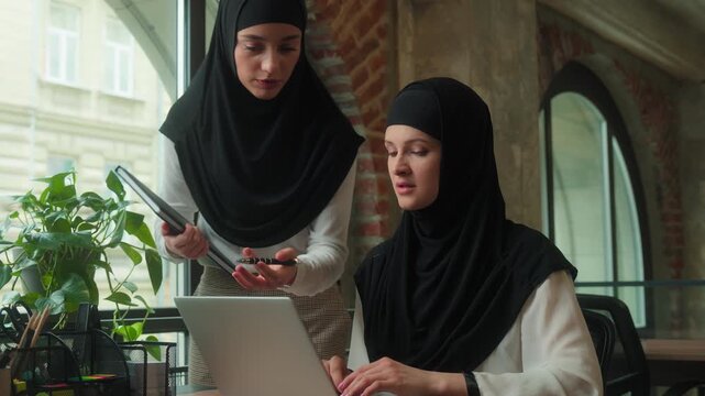 Two colleagues team business women in hijab girls Islamic female intern working laptop computer corporate software using serious arabian leader mentor coming talking explain holding document in office