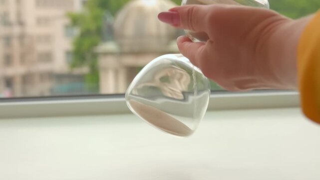 A female hand flips a glass hourglass on a stand by a window. Time flows like sand. Visual metaphor for time is money and the passage of time