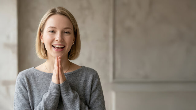 Happy blonde woman in grey sweater gesturing namaste or thank you with hands together and copy space.