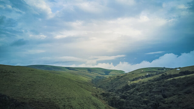 Aerial view of lush, velvety green hills rolling into a deep, shadowy valley beneath a dramatic, cloud-strewn sky, Obudu, Cross River, Nigeria.