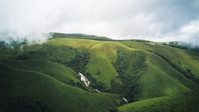 Aerial view of verdant rolling hills with a cascading white waterfall carving through the lush green landscape under a dramatic cloudy sky, Obudu, Cross River, Nigeria.