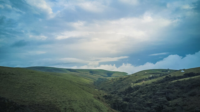 Aerial view of rolling green hills and valleys bathed in the dramatic light of a cloudy sky, with subtle hints of distant structures, Obudu, Cross River, Nigeria.