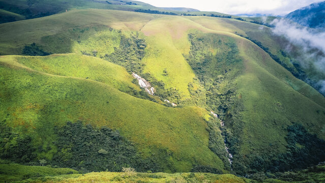 Aerial view of vibrant green rolling hills with a sparkling waterfall cascading through a rocky gully, kissed by soft clouds, Obudu, Cross River, Nigeria.