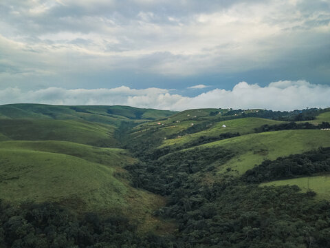 Aerial view of lush, emerald green rolling hills embracing a deep, shadowy valley of dense forest under a dramatic, cloud-strewn sky, Obudu, Cross River, Nigeria.