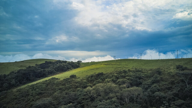 Aerial view of rolling emerald hills crowned with a dense, dark green canopy under a dramatic expanse of white and deep blue clouds, Obudu, Cross River, Nigeria.