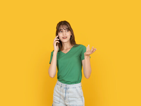 Young woman talking on smartphone with expressive hand gesture, reacting during conversation with curious and engaged expression. Isolated on bright yellow background with copy space.
