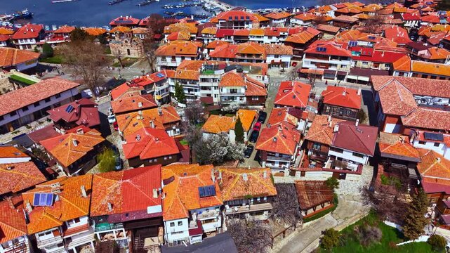 Houses with orange roofs form a tight layout in Nesebar Old Town. The scene captures the buildings near the Black Sea with boats visible in the water.