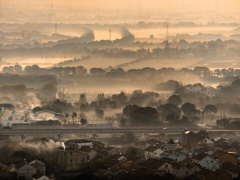 Aerial view of a town's warm-toned roofs and an elevated road piercing through layers of ethereal mist, all bathed in a soft golden glow, Hu Zhou Shi, Zhe Jiang Sheng, China.