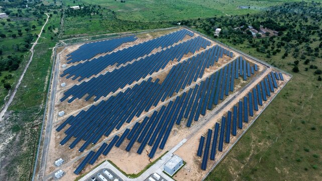 Aerial view of a vast solar farm with rows of dark, gleaming panels starkly contrasting against the reddish-brown earth and lush green landscape, Maiduguri, Borno, Nigeria.