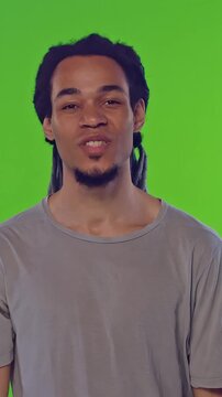 Young African American man with dreadlocks smiles warmly at the camera, standing on green screen background. His relaxed posture and friendly expression convey confidence, positivity, and openness