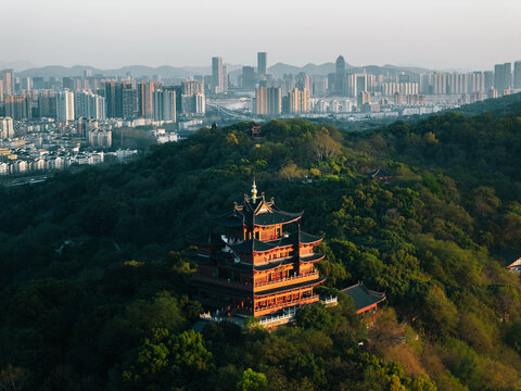 Aerial view of a striking traditional pagoda, its dark wood and crimson details vibrant against lush green hills, overlooking a modern city skyline, Hang Zhou Shi, Zhe Jiang Sheng, China.
