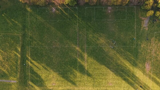 Aerial top-down view of two grass soccer fields in a public park at sunset, people gathering for a match, England.