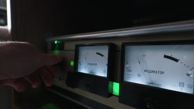 A close-up view of a hand turning the volume knob on a vintage analog audio amplifier. The lit retro dials feature Russian text translated as Indicator. Green lights glow under the meters.