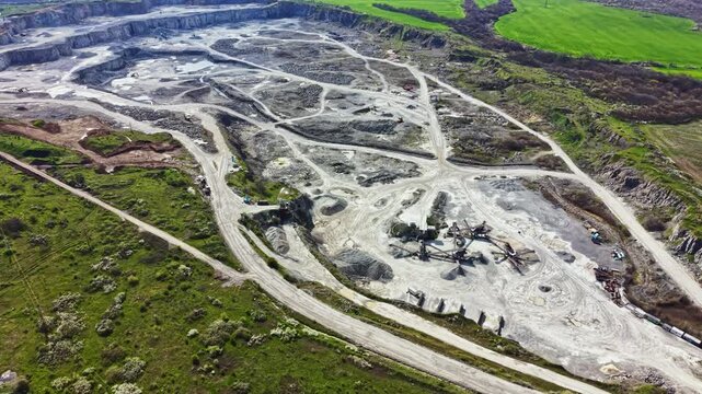 Large granite quarry shows multiple machinery and paths where workers extract granite. The area features open ground and nearby green fields under clear skies during daylight.