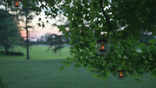 Glass mason jar lanterns with warm flickering candles hang from a leafy green tree branch at dusk, overlooking a peaceful, expansive lawn during a summer sunset.