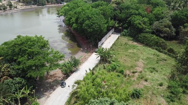 long, narrow road paved on the rugged terrain of a forested agricultural settlement at the outskirts of a small town. Drone, aerial