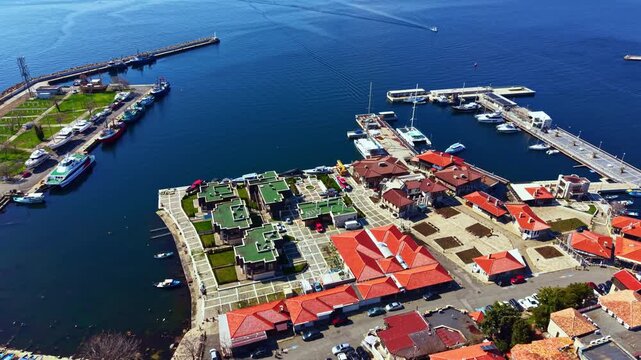 Visitors walk through Nesebar Old Town near the marina where boats are docked. The scene shows buildings and open spaces under bright sunny weather.