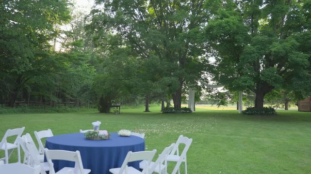A serene wide shot of an outdoor wedding venue at sunset, featuring a blue-clothed table and white chairs on a manicured lawn, with an elegant draped archway in the background.