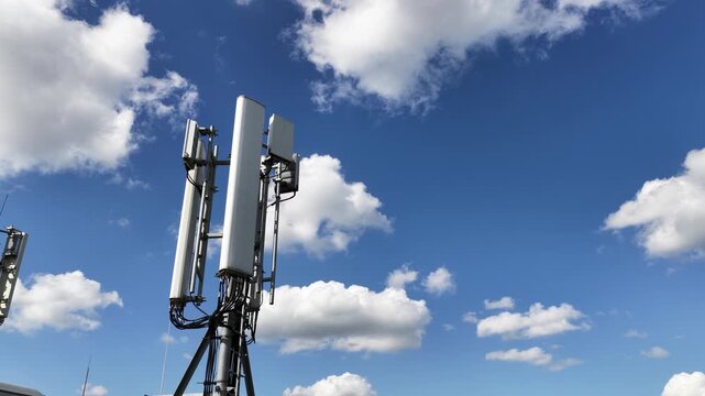 Telecommunication tower with 5g and 4g antennas providing mobile network coverage, with white clouds moving across a bright blue sky, representing modern wireless connection technology