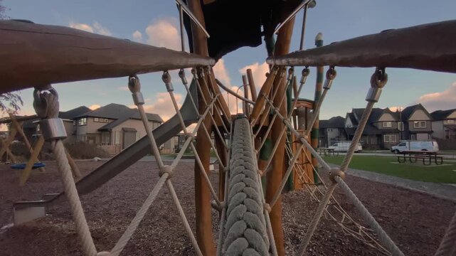 Thick woven climbing rope stretching across timber play structure hanging above wood chips framed against suburban housing beneath cloudy sunset sky captured via backward moving viewpoint
