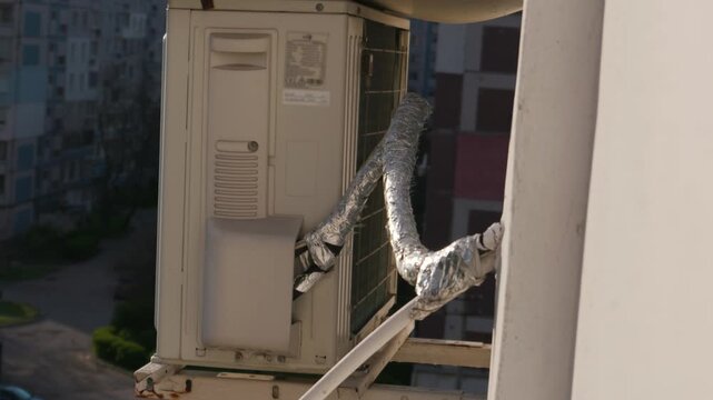 An outdoor air conditioning unit is mounted on a building wall with insulation pipes wrapped in silver foil. The external AC compressor system shows signs of wear and maintenance in the city.