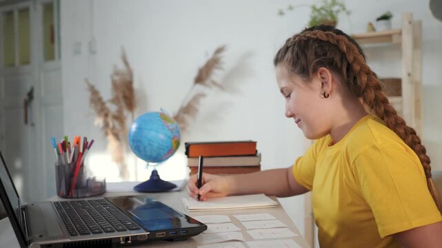 Studying girl arranging flashcard while writing homework at desk with laptop and globe child student focused on study and learning with book stack and notebook in home school education setting
