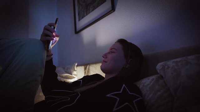 Girl doom scrolling her smartphone on a coach at night. Social network and phone addiction concept. Cozy home ambient light