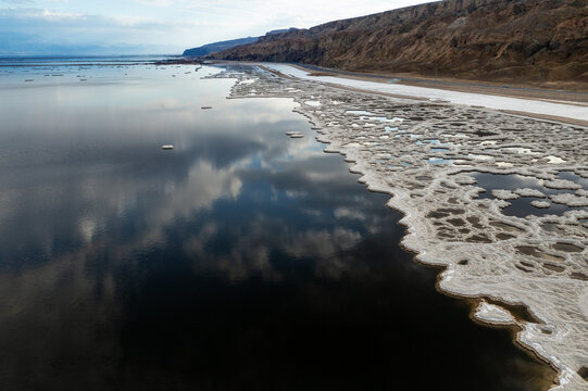 Aerial view of ice floes scattered across the dark, reflective waters near a rugged shoreline, contrasting sharply with the pale sky, Israel.